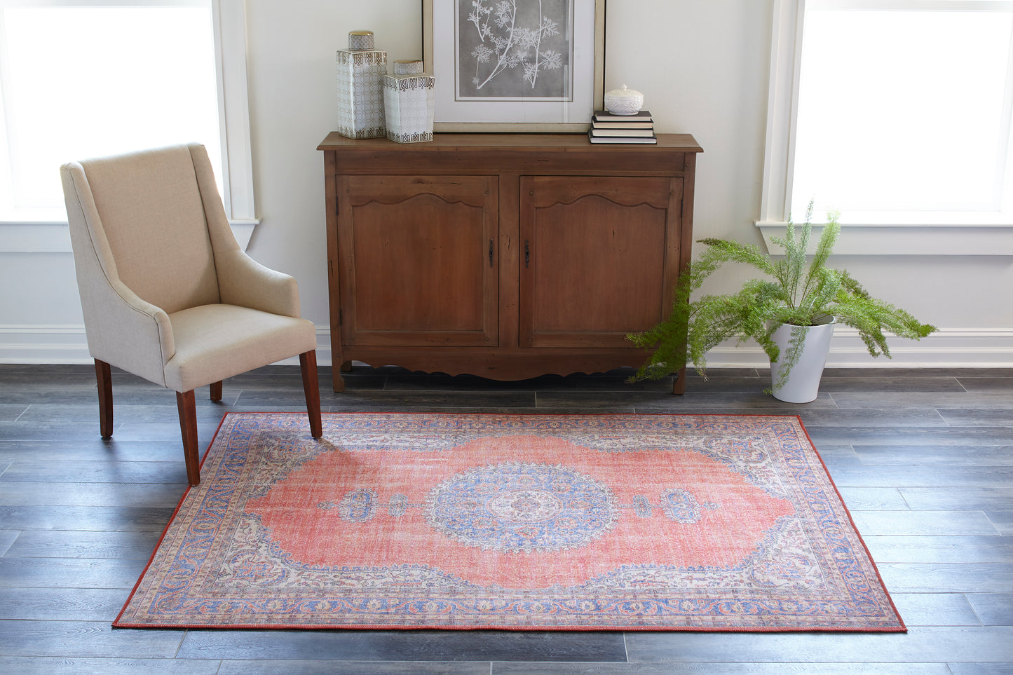 Coral and blue ornate area rug in a living room with beige upholstered wingback chair, wooden sideboard, and potted fern.