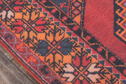 Red oriental rug with orange, navy & ivory geometric floral motifs & bordered edge on a wooden floor.