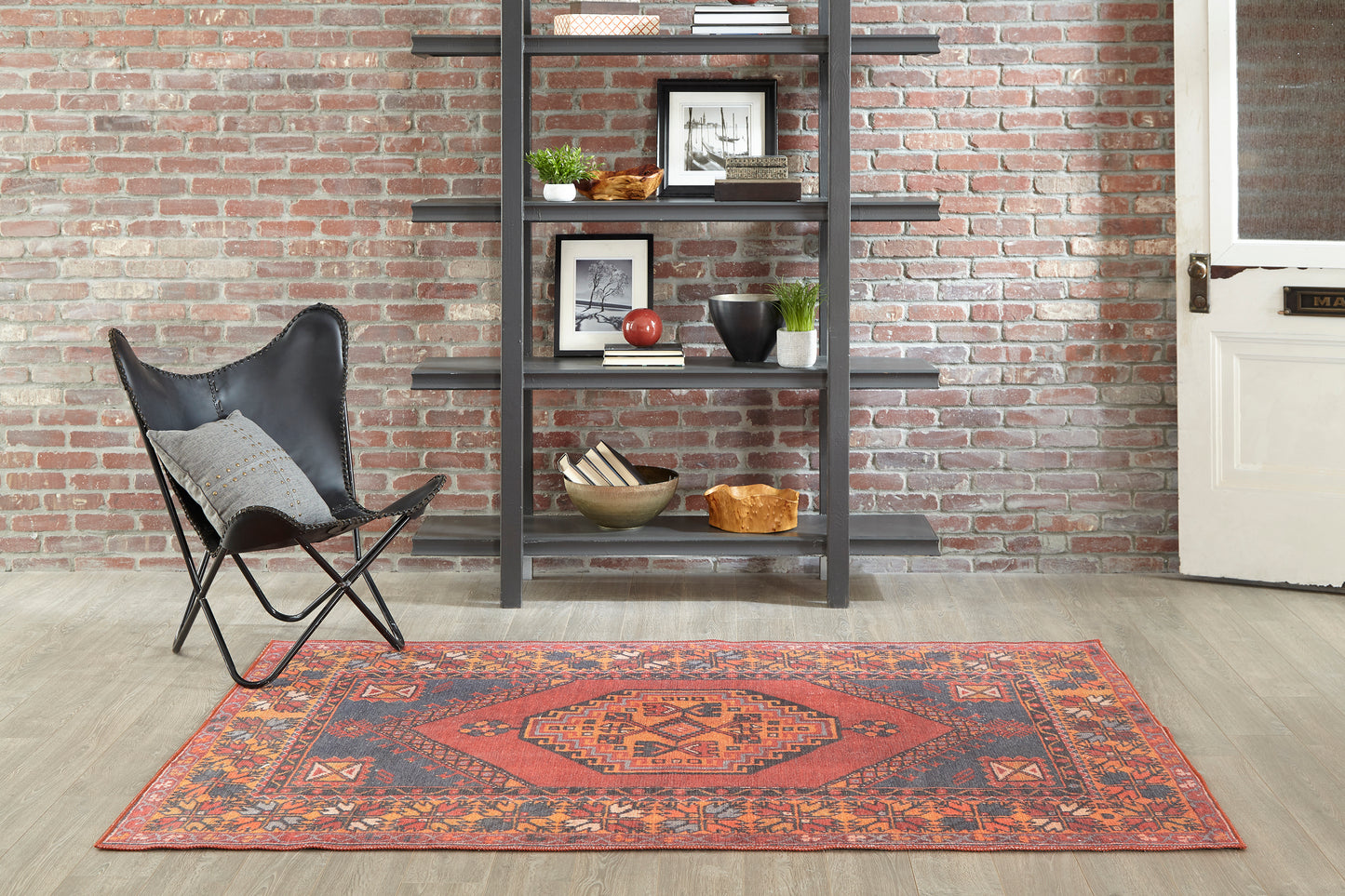 Red geometric oriental rug with blue border on light wood floor beside a black metal ladder shelf & a black butterfly chair.