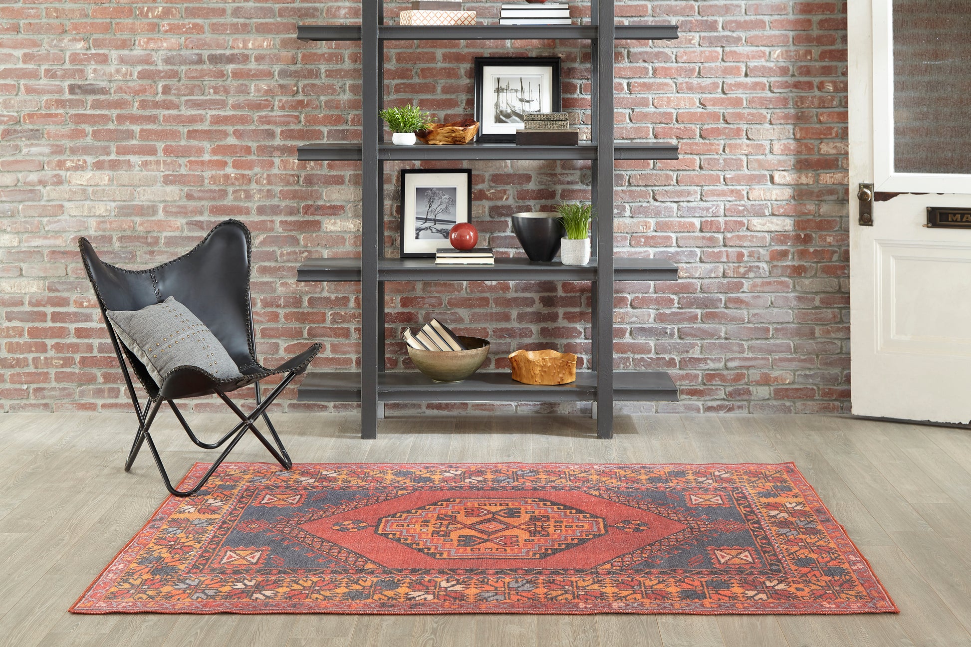 Red-orange geometric oriental rug on light wood floor beside a black metal bookshelf and leather butterfly chair.