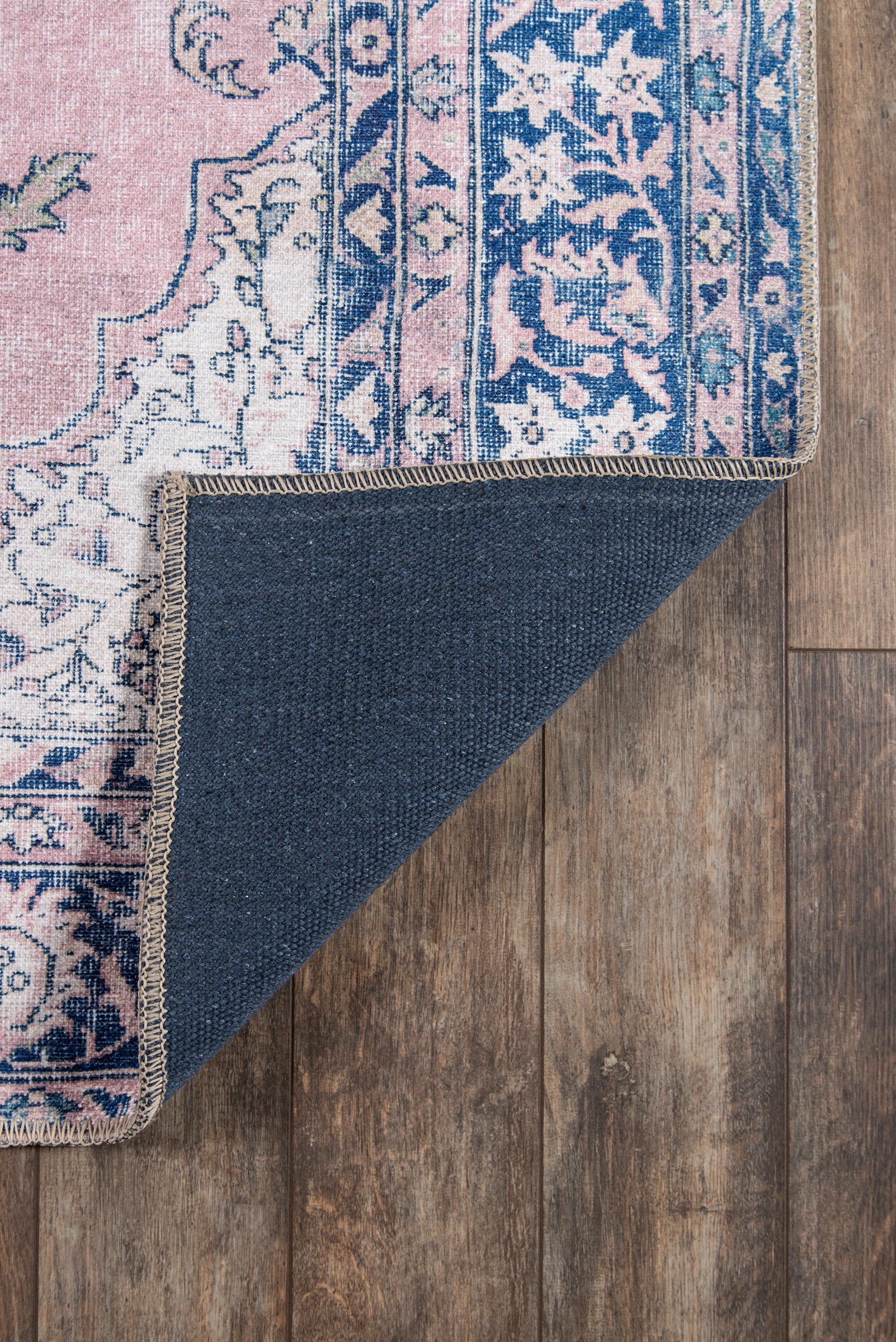 Pink & blue floral patterned area rug with fringe border; folded corner reveals navy underside on wooden floor.