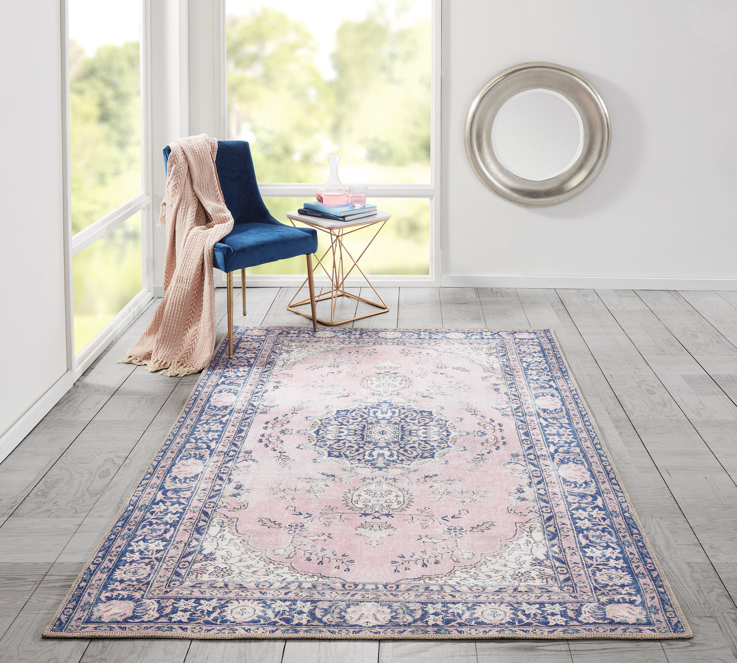 Pink center rug with a blue ornate border in a bright living space; blue velvet chair, blush throw, and gold-glass side table.