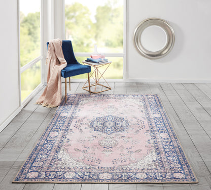 Pink & blue rectangular Persian-style area rug with ornate border in a bright room, blue velvet chair & beige throw.