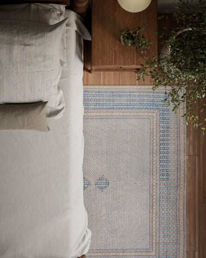 Top-down view of a cozy bedroom: beige bed linens, wooden nightstand, blue-beige patterned runner rug, and a green plant.