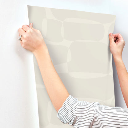 Two hands in striped sleeves hold a large beige wallpaper sheet with a soft rounded-oval pattern.