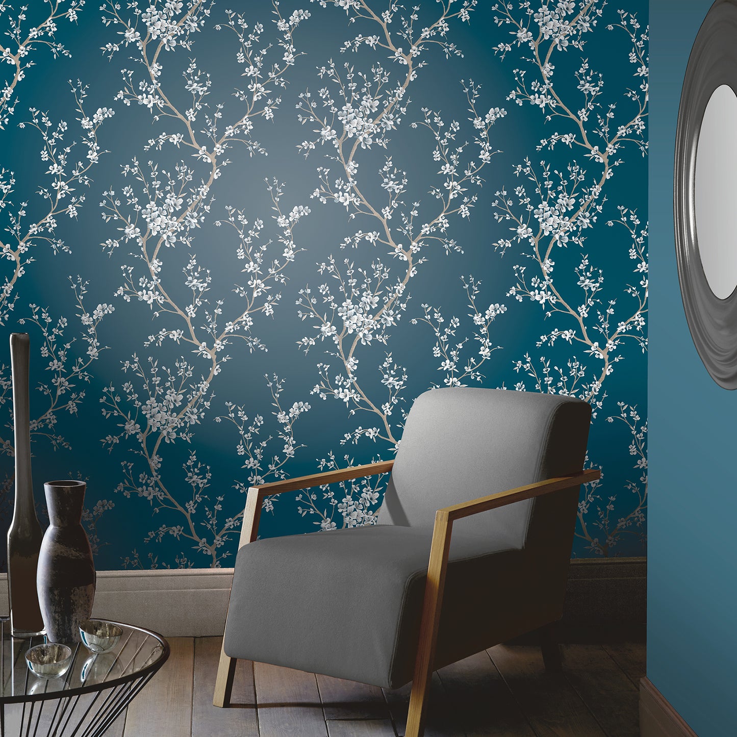 Blue flowering tree wallpaper with a botanical motif; gray armchair with wooden frame, glass coffee table & vases.