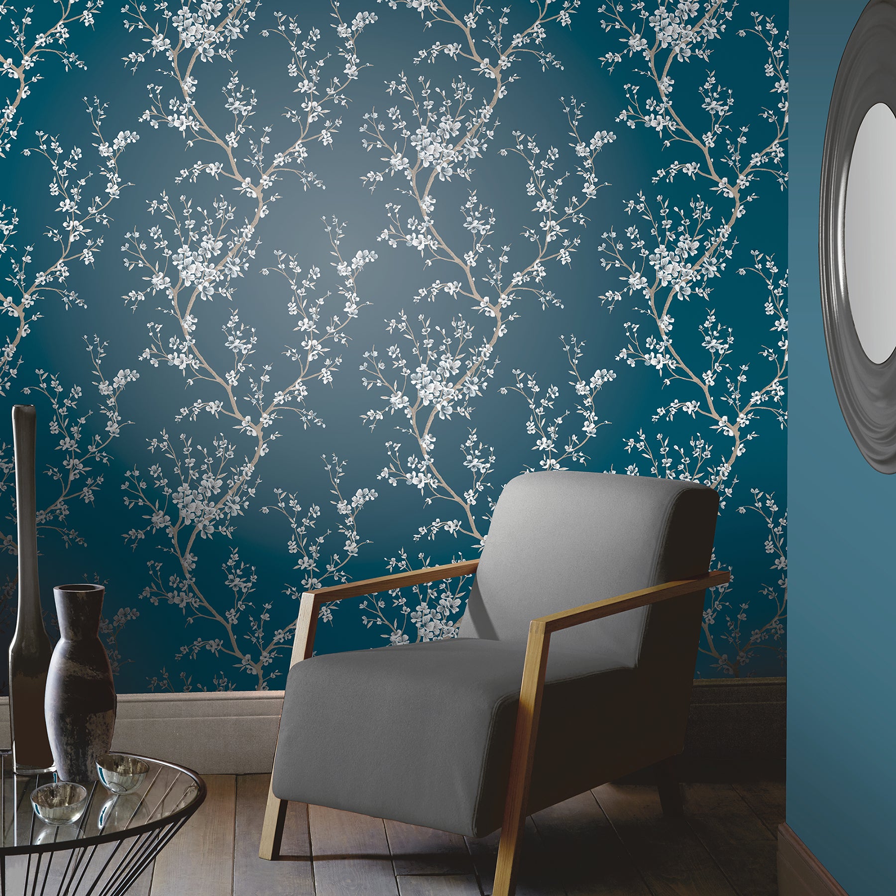 Blue flowering tree wallpaper with a botanical motif; gray armchair with wooden frame, glass coffee table & vases.