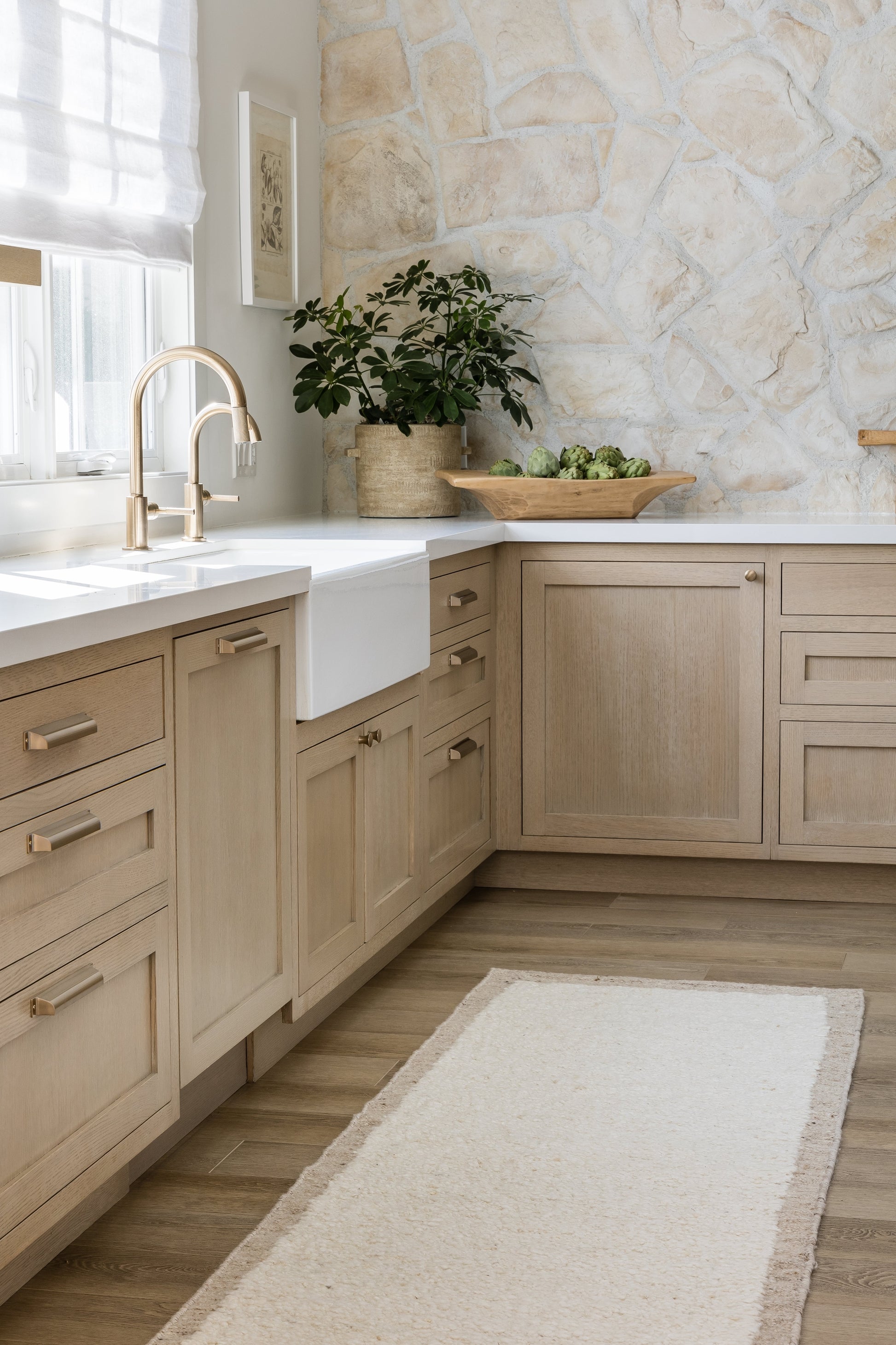 Beige kitchen with light wood cabinets, white farmhouse sink, brass faucet, stone wall, potted green plant, and beige rug.