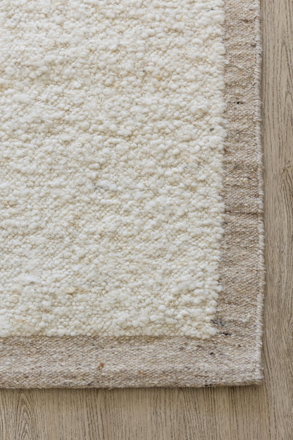 Cream shag rug with beige border on light wood plank floor; close-up shows fluffy tufted texture.