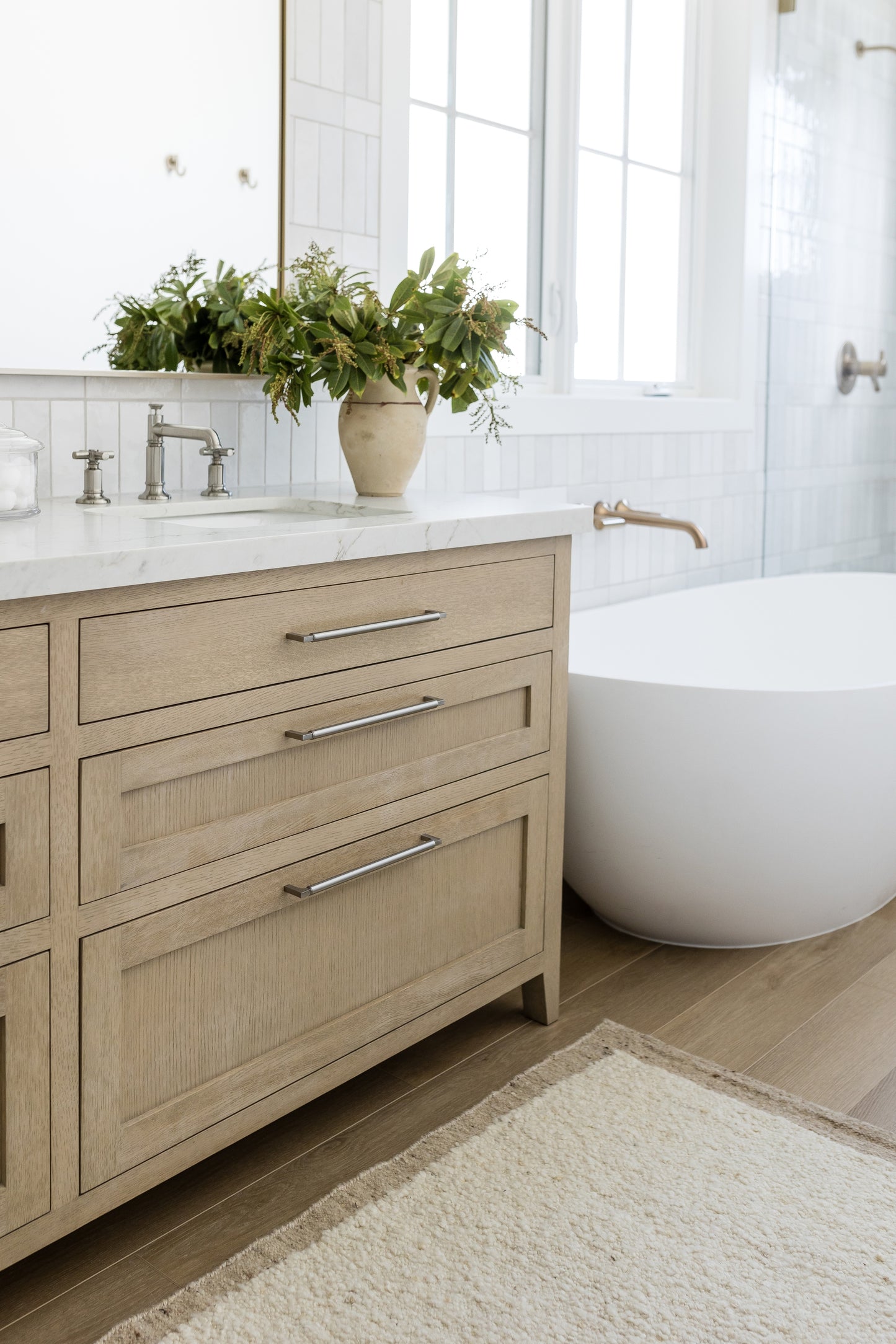 Beige wood vanity with three drawers, chrome handles, marble top; potted greenery, freestanding white tub, white tiles.