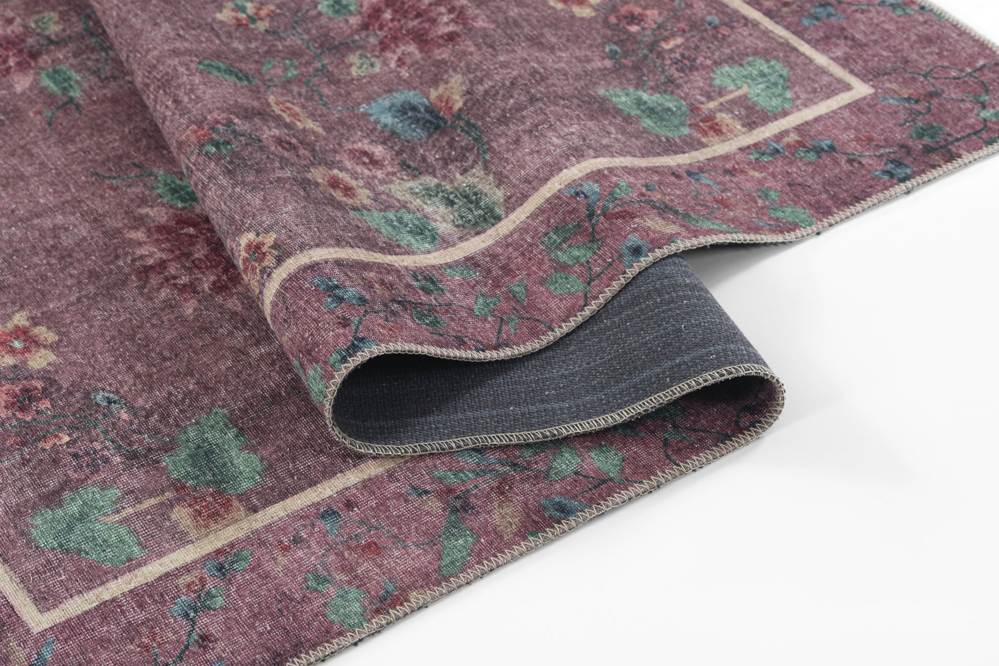 Purple rug with green floral motifs and a cream border; rolled edge showing grey backing.