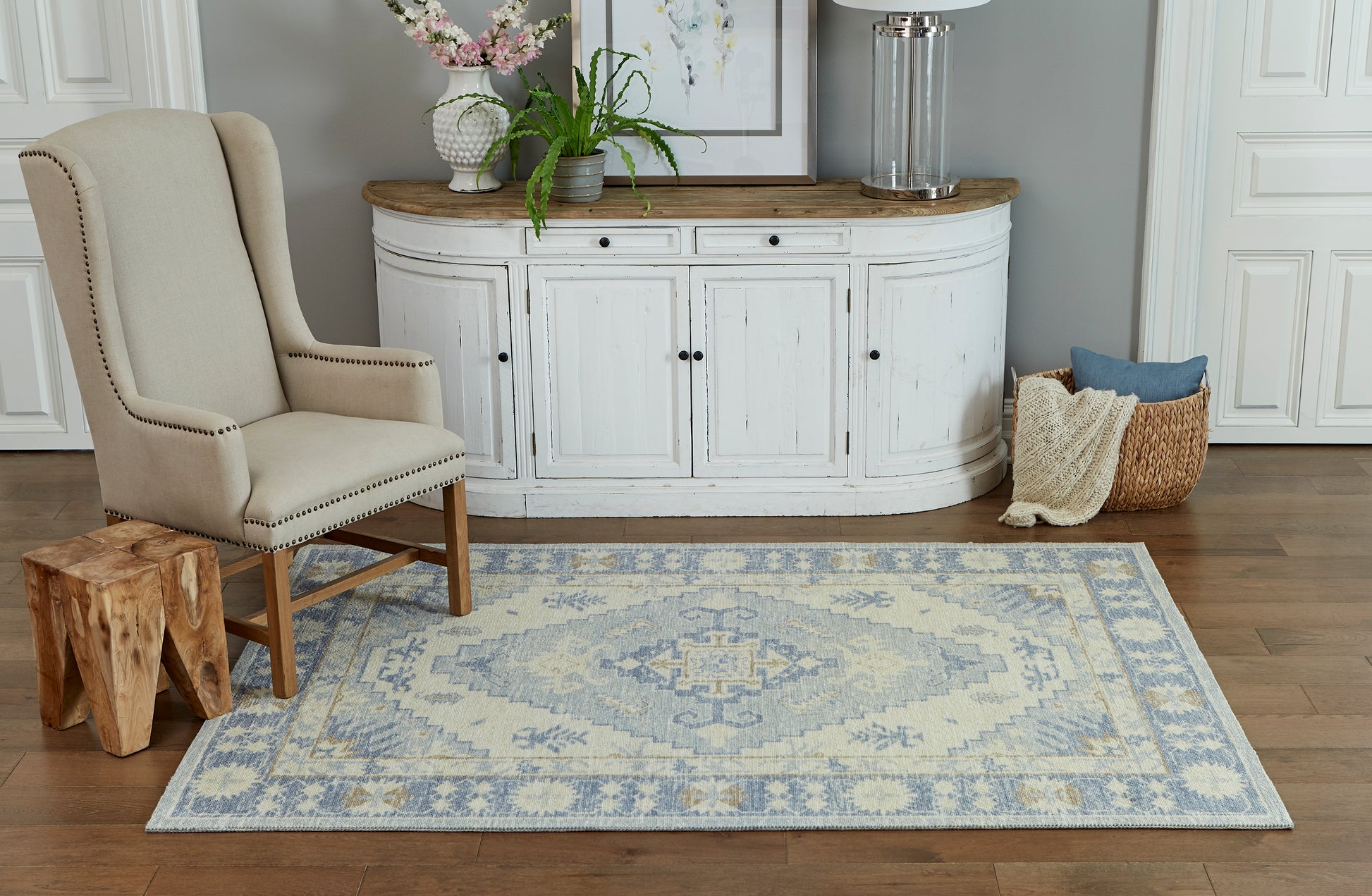 Beige upholstered wingback chair beside curved white console with wood top, blue patterned rug, potted plants, woven basket.