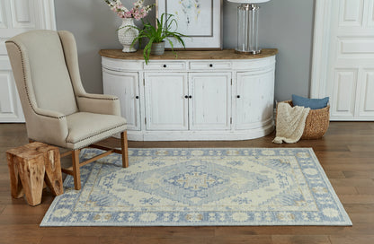 Beige upholstered wingback chair beside curved white console with wood top, blue patterned rug, potted plants, woven basket.