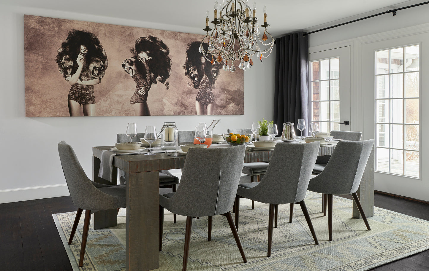 Dining room with a long wooden table, gray upholstered chairs, crystal chandelier, triptych art, rug, and French doors.