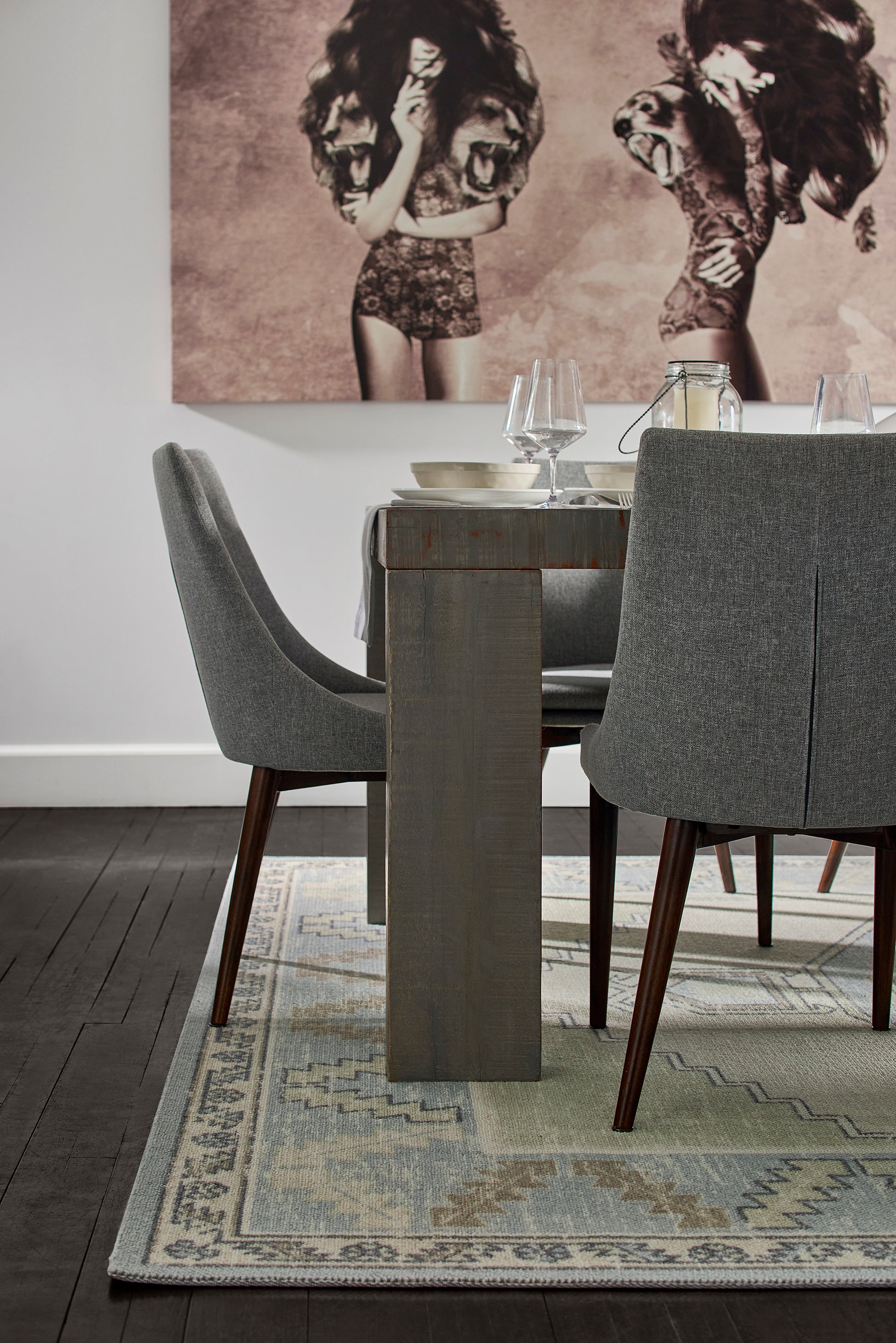 Dining room with grey upholstered chairs, rustic wood pedestal table, blue-gray rug, and sepia art of dancing figures.