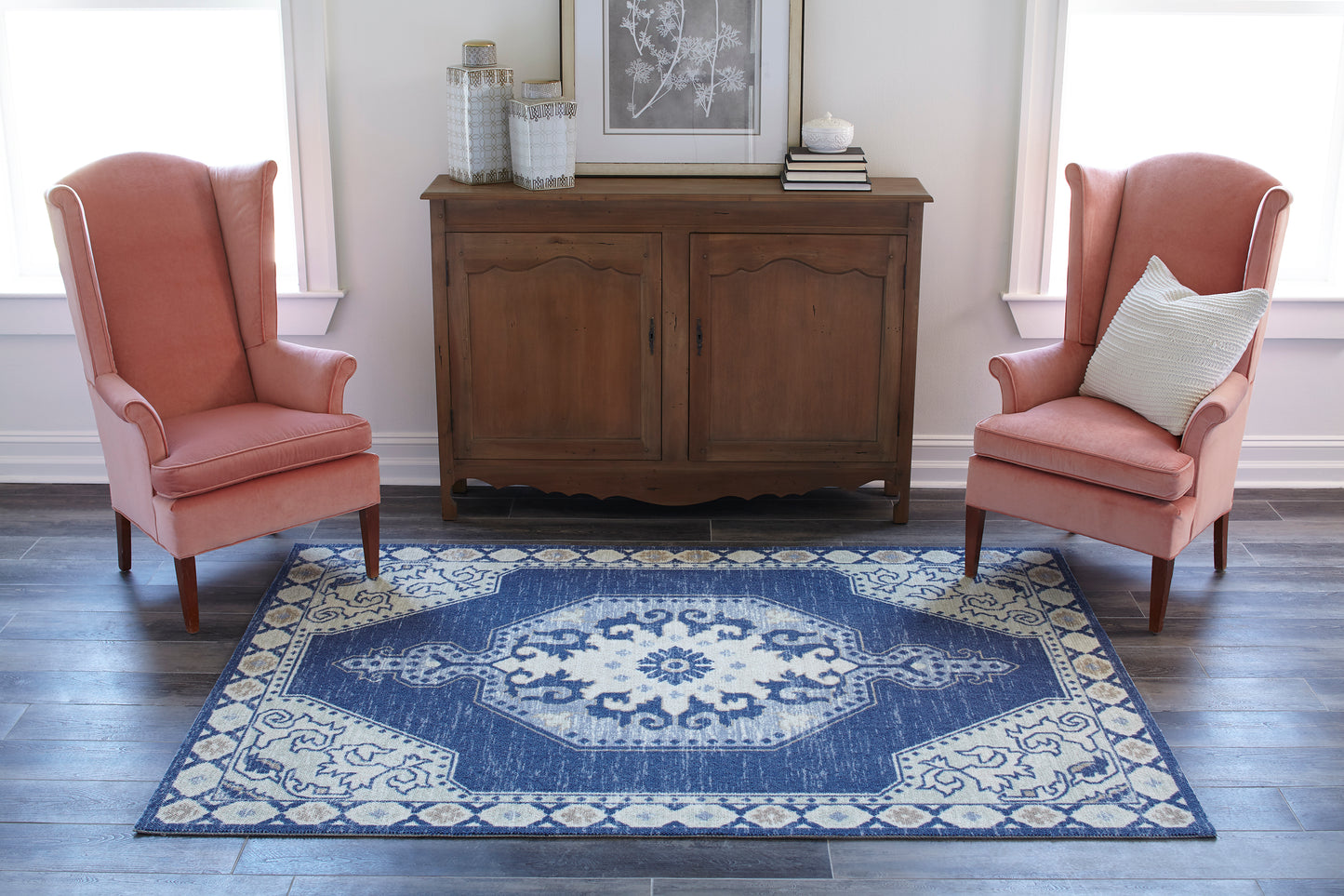 Two coral pink wingback chairs flank a wooden sideboard atop a blue and cream ornate area rug.
