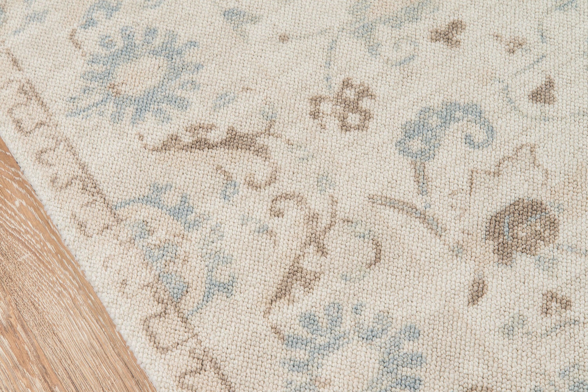 Rectangular cream woven rug with pale blue and brown floral pattern, close-up on wooden floor edge.