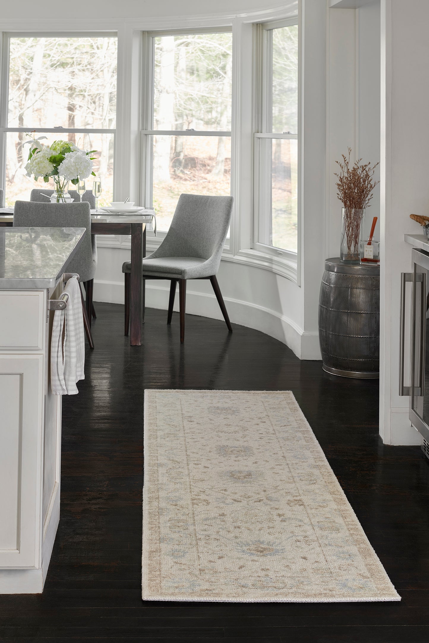 Beige patterned runner rug on dark hardwood floor in a bright white kitchen with a dining table and gray chairs by curved bay windows.