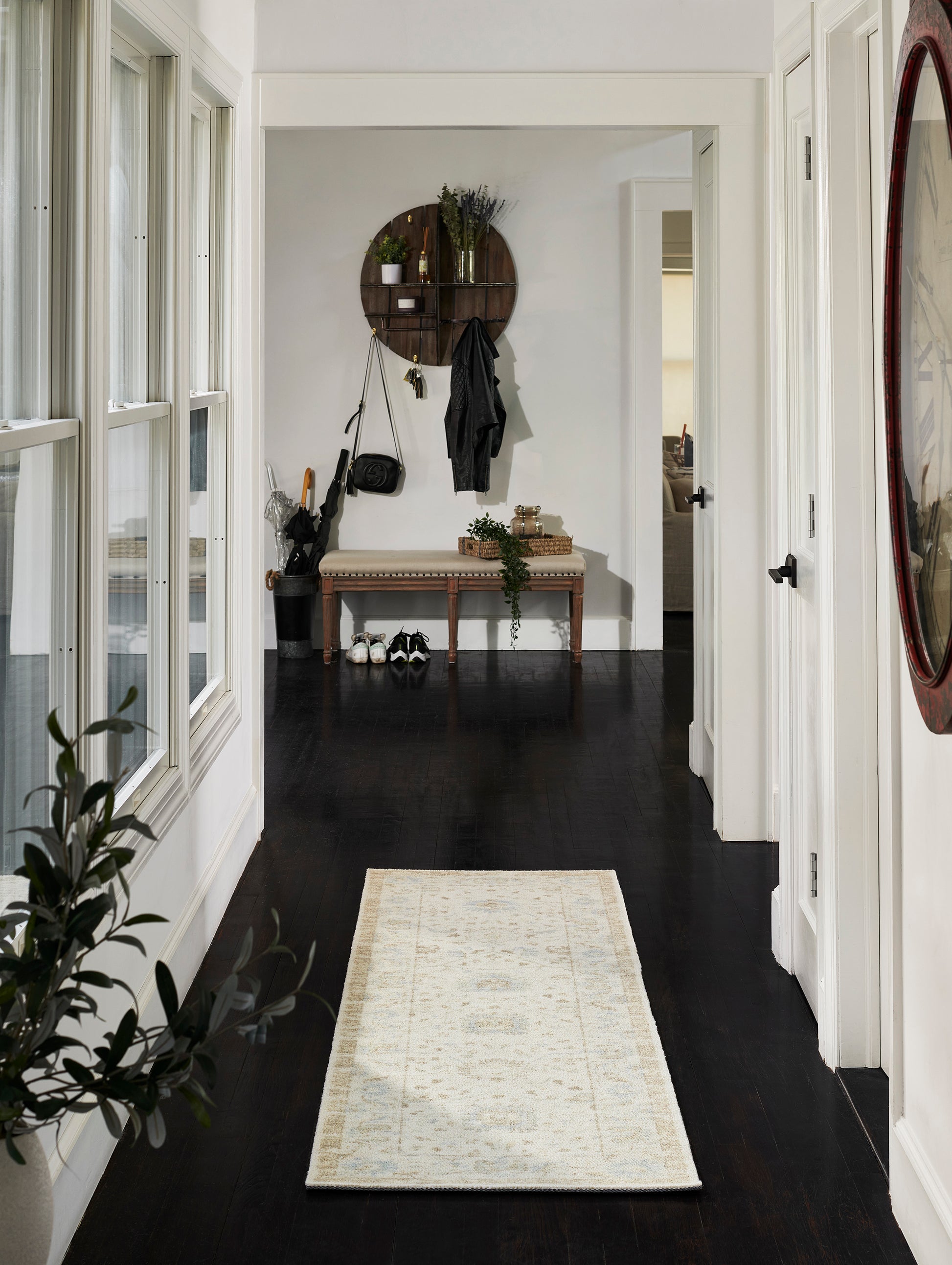 Bright entry hall with dark hardwood floor, beige runner rug, white walls; wooden wall decor, bench, hooks, plants, coats.