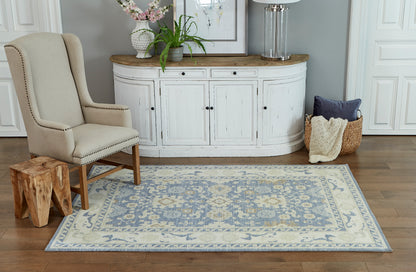 Beige wingback chair, wood legs, natural stump side table, blue ornate rug, curved white cabinet, plants and lamp.