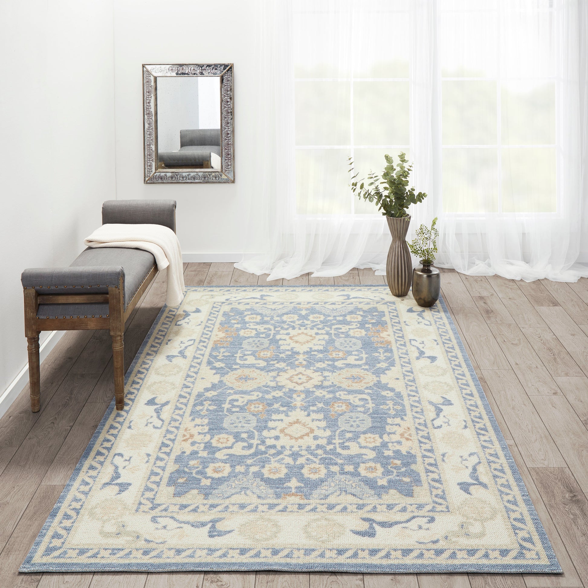 Blue-beige ornate area rug on wooden floor beside a gray upholstered bench; wall mirror, sheer curtains, and botanical vases.