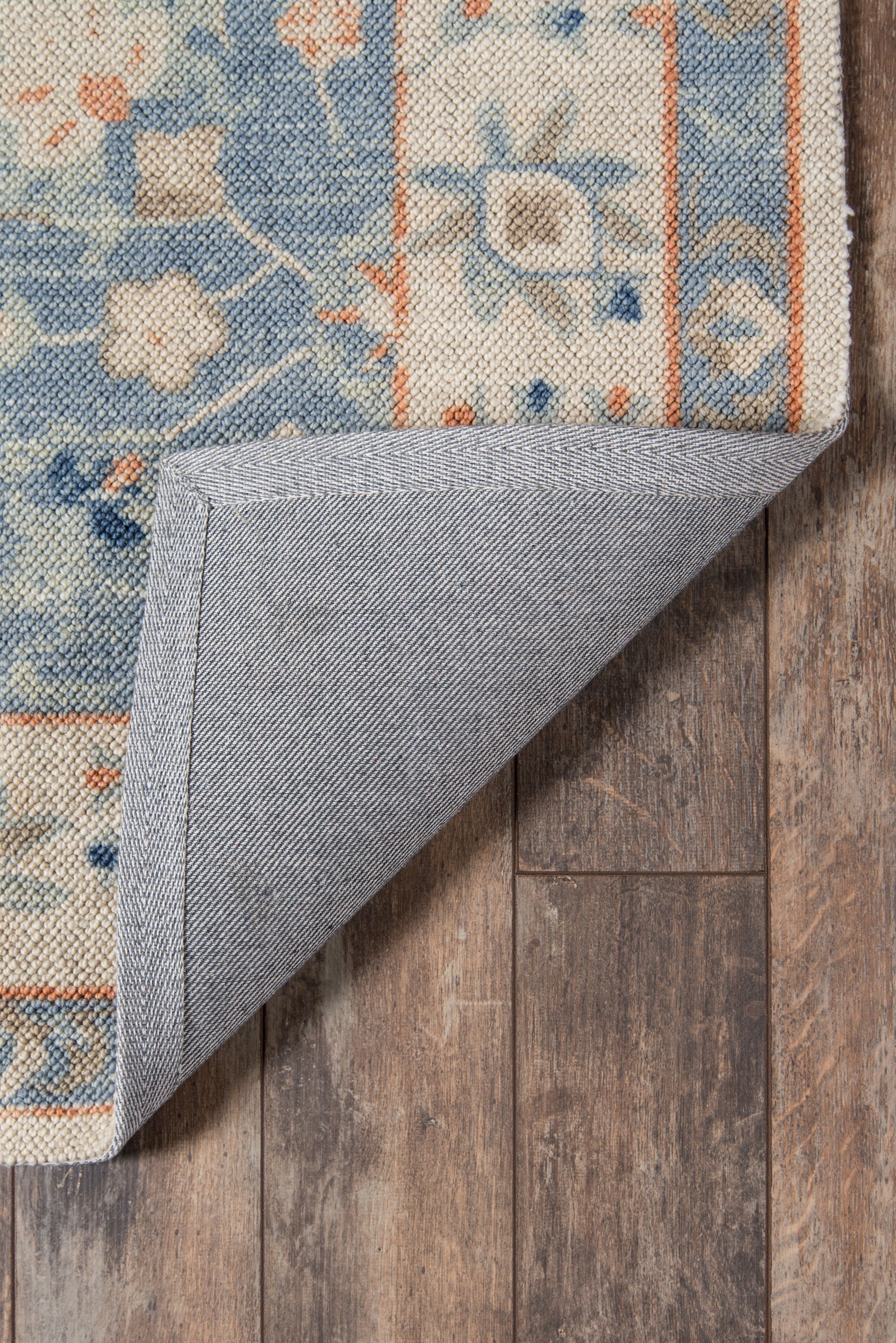 Blue floral patterned rug with cream & orange accents; folded corner reveals grey backing on wooden floor.