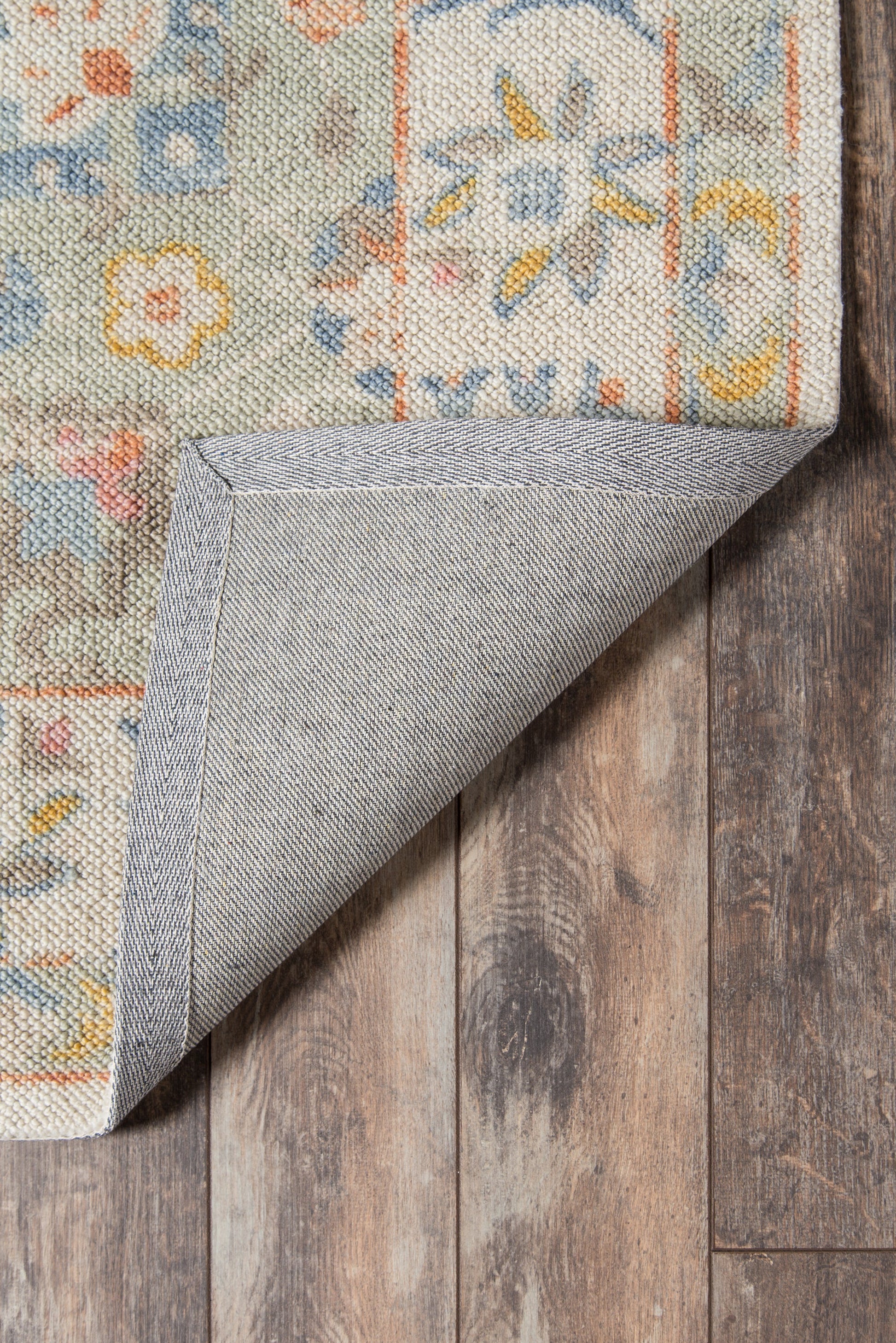 Pastel floral woven rug with blue, orange and yellow hues; folded corner reveals gray backing on a wooden floor.
