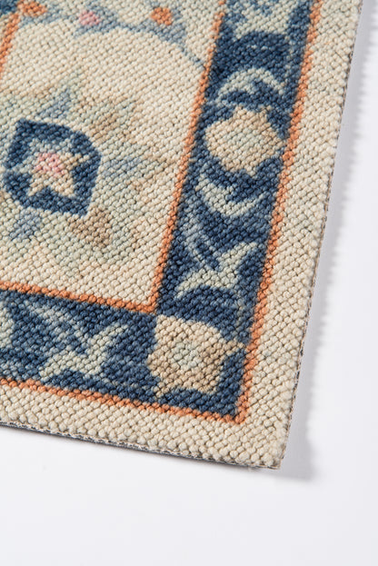 Anatolian-style woven rug corner: cream field, blue border with orange trim, floral motifs, chunky knot texture.