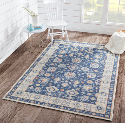Blue oriental-pattern area rug with cream border and floral motifs on a wooden floor beside a white chair.