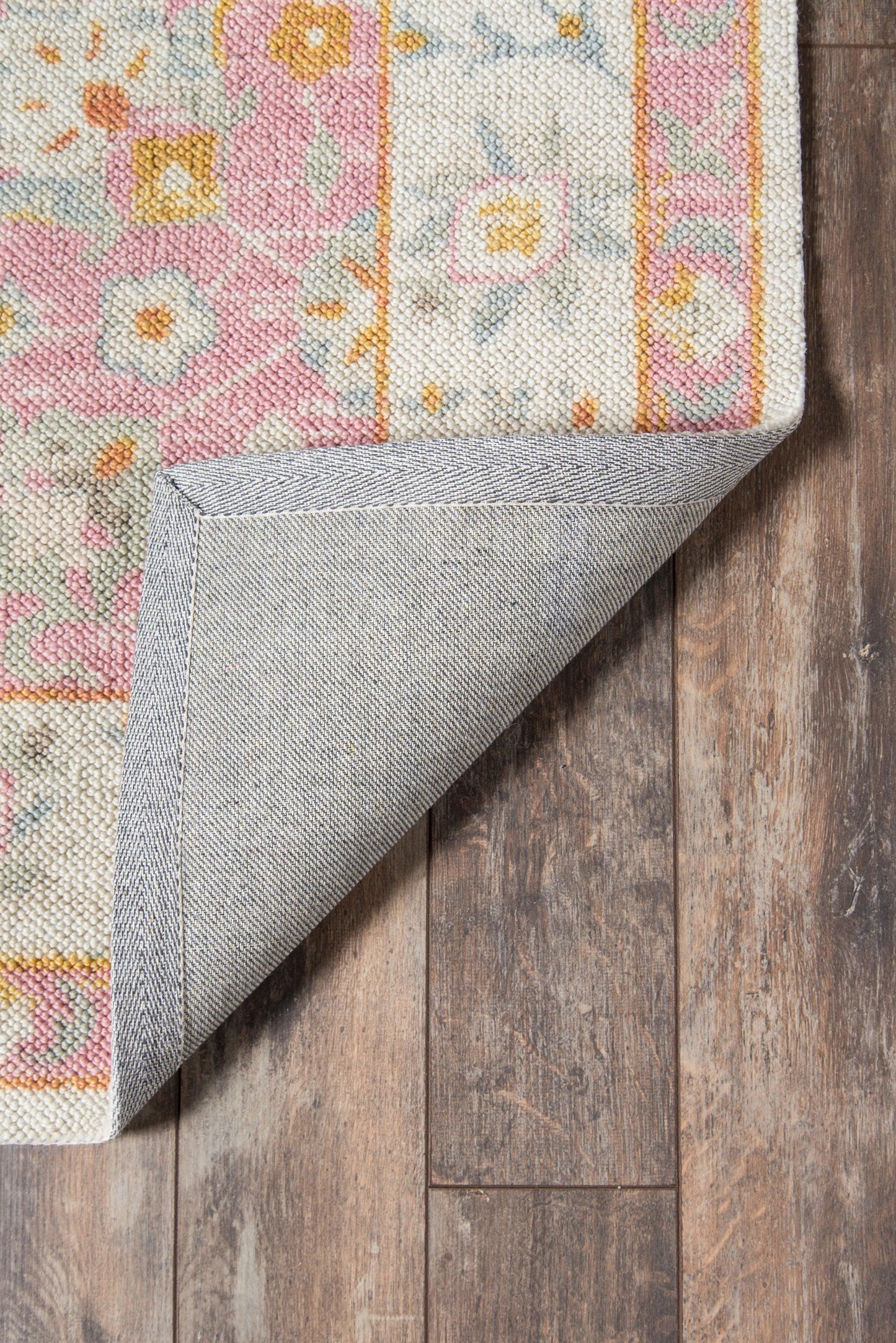 Pink and ivory patterned area rug with orange accents, folded corner reveals gray backing on wooden floor.