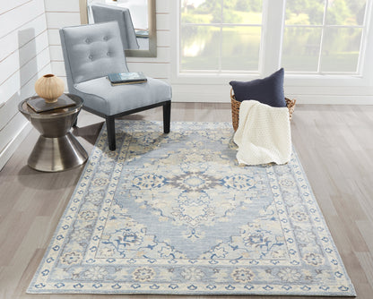 Blue-gray ornamental area rug with a beige border in a bright living room; tufted gray chair, a metal side table, and a basket with a cream throw.
