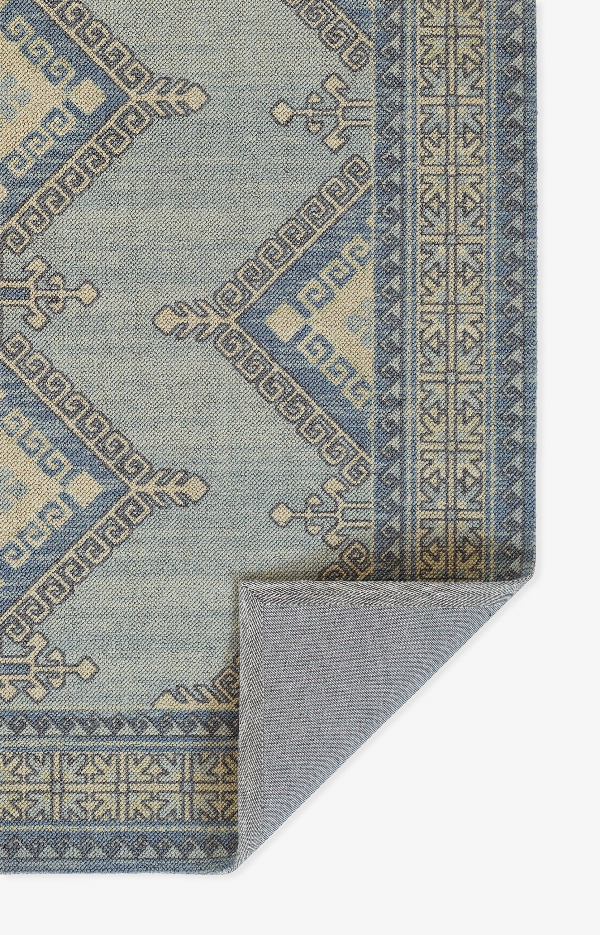 Blue-gray rectangular woven rug with geometric lattice and Greek key border, folded corner revealing gray backing.