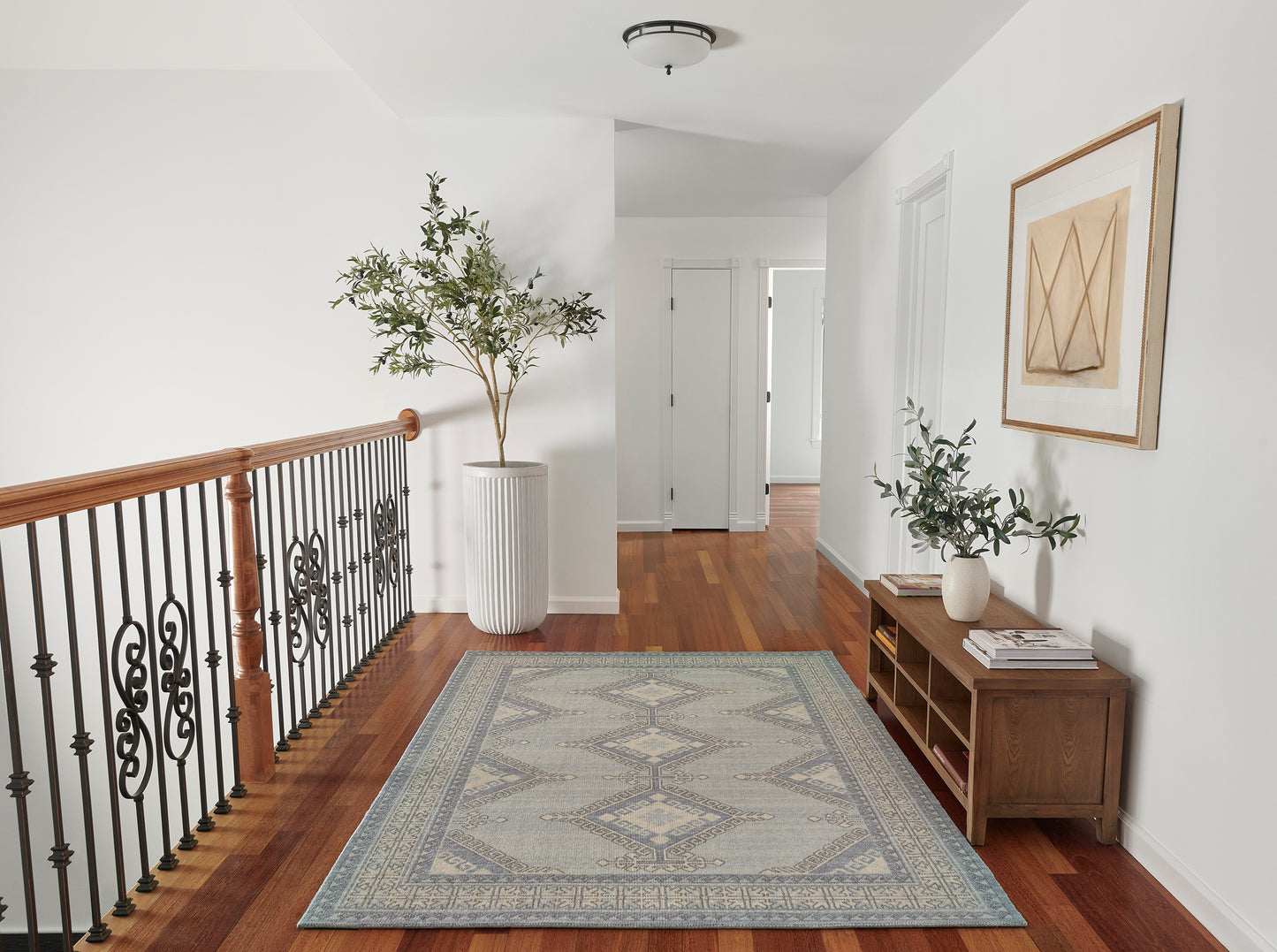 Blue-gray geometric area rug on hardwood floor beside a wooden handrail with iron balusters, tall plant, console table, and framed art.