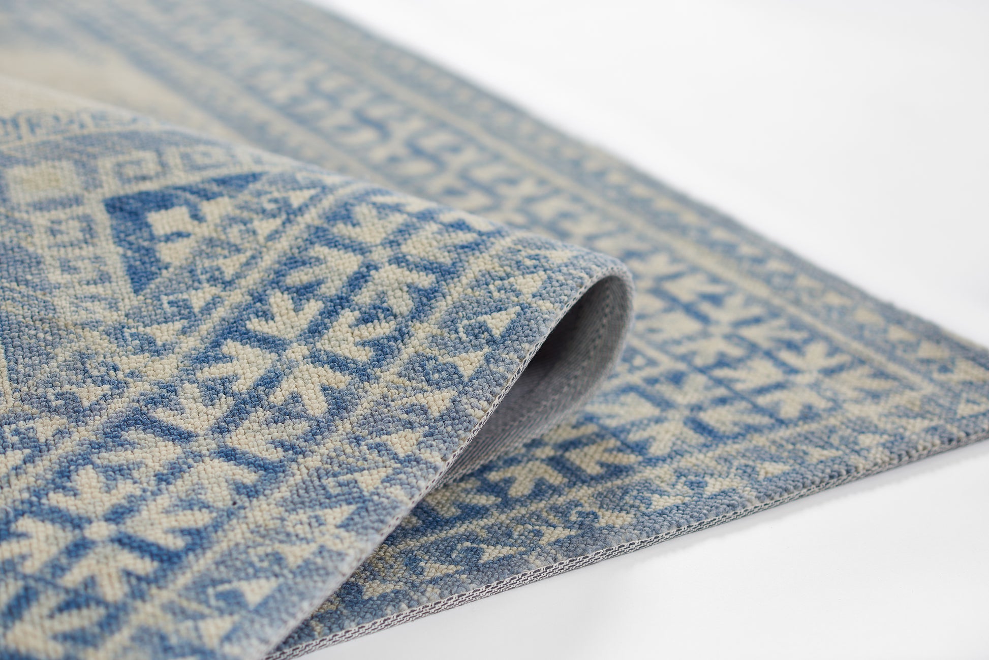Blue and ivory woven wool rug with geometric diamond motifs, rolled edge showing soft textured weave.