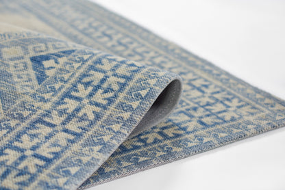 Blue and ivory woven wool rug with geometric diamond motifs, rolled edge showing soft textured weave.