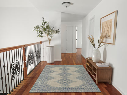 Bright entryway with hardwood floors, blue geometric rug, wooden console with white vase of dried grasses, framed art, potted tree & iron railing.