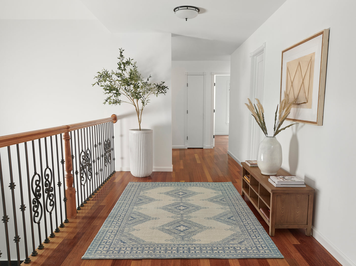 Bright hallway landing with hardwood floors, black wrought-iron railing, tall potted tree, blue geometric rug, wooden console & vase.