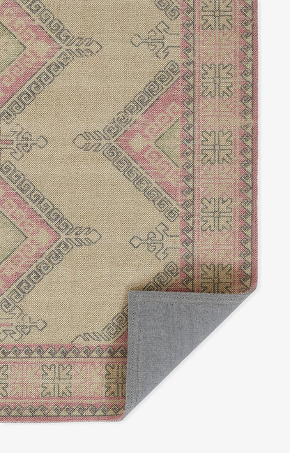 Beige woven rug with pink geometric diamonds and olive border; pink accents, with gray underside visible at corner.
