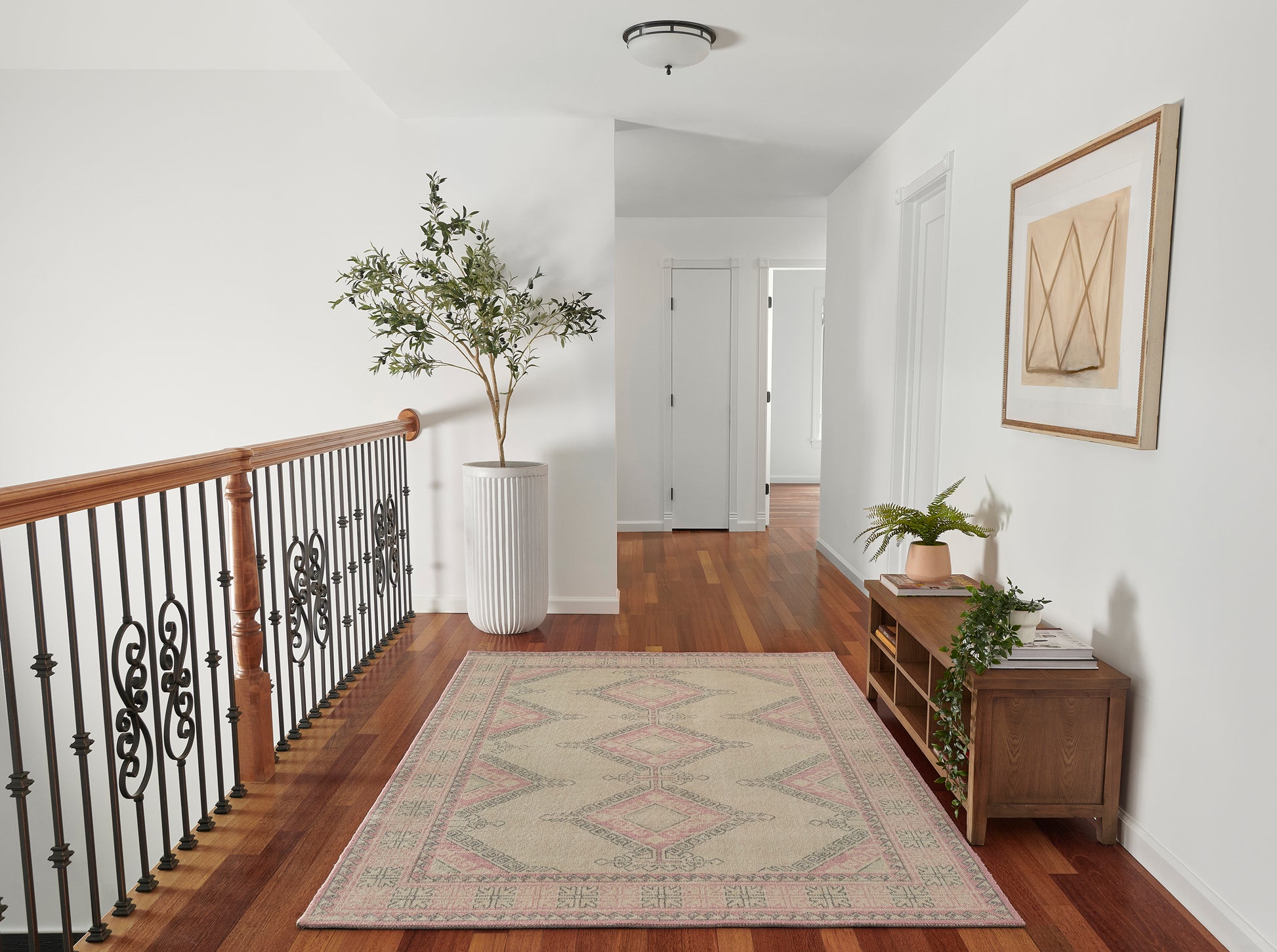 Hallway with pink geometric rug, hardwood floor, iron-and-wood railing, tall potted plant, console with books, abstract art.