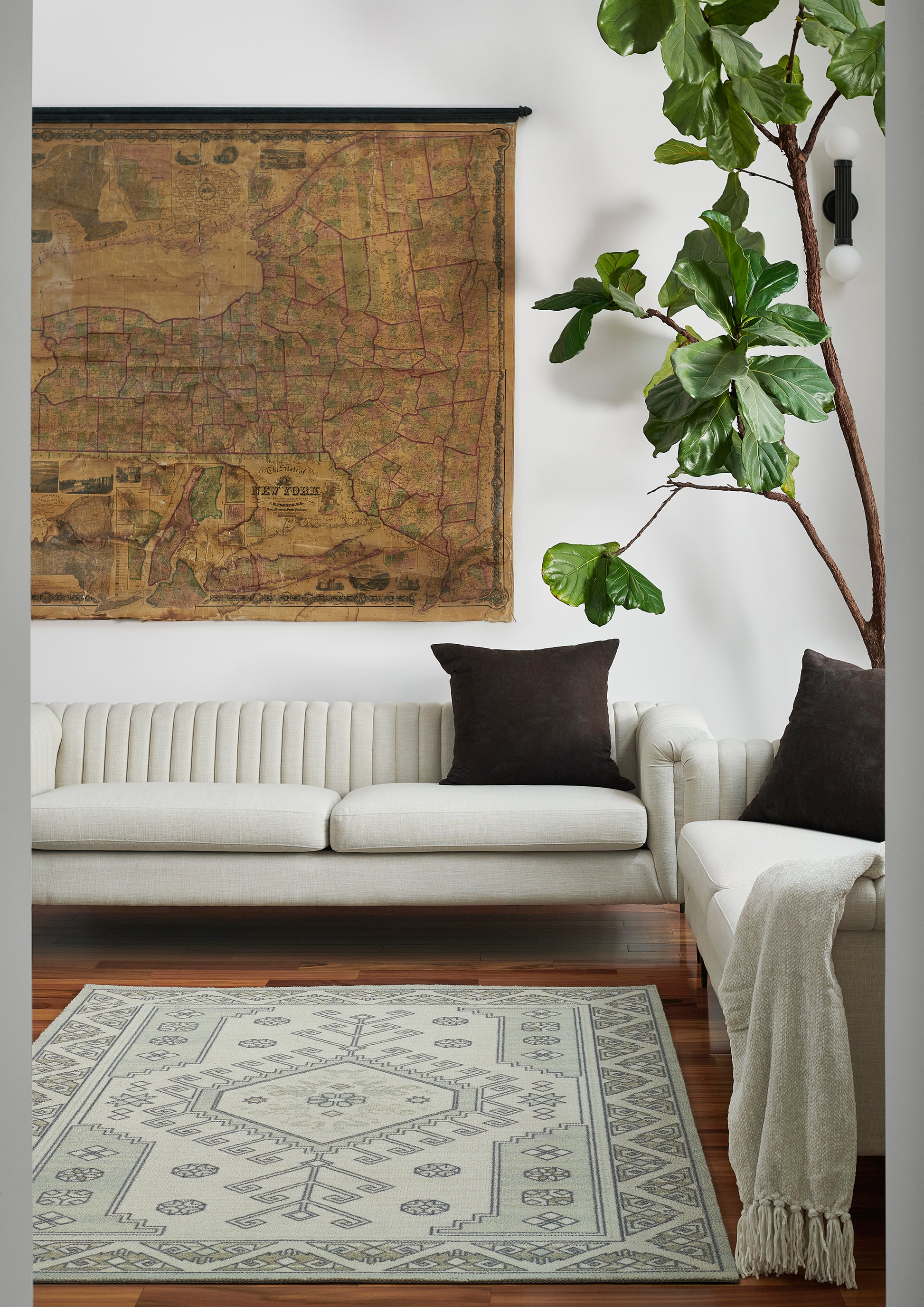 Living room with vintage map wall art, cream sofa with brown pillows, tall plant, and a grey geometric-pattern rug.
