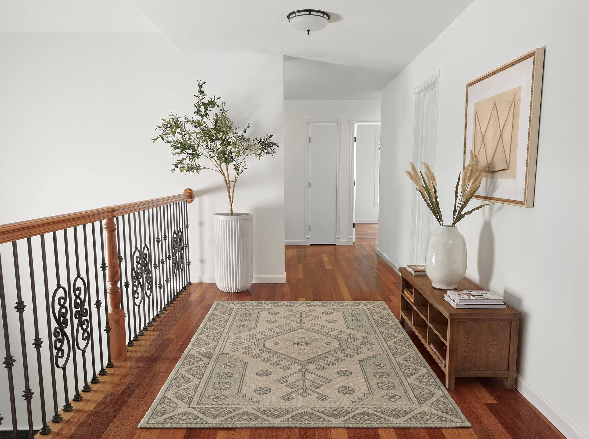 Bright hallway with hardwood floor, beige patterned rug, potted plant, wrought-iron railing, wooden console with vase & stems