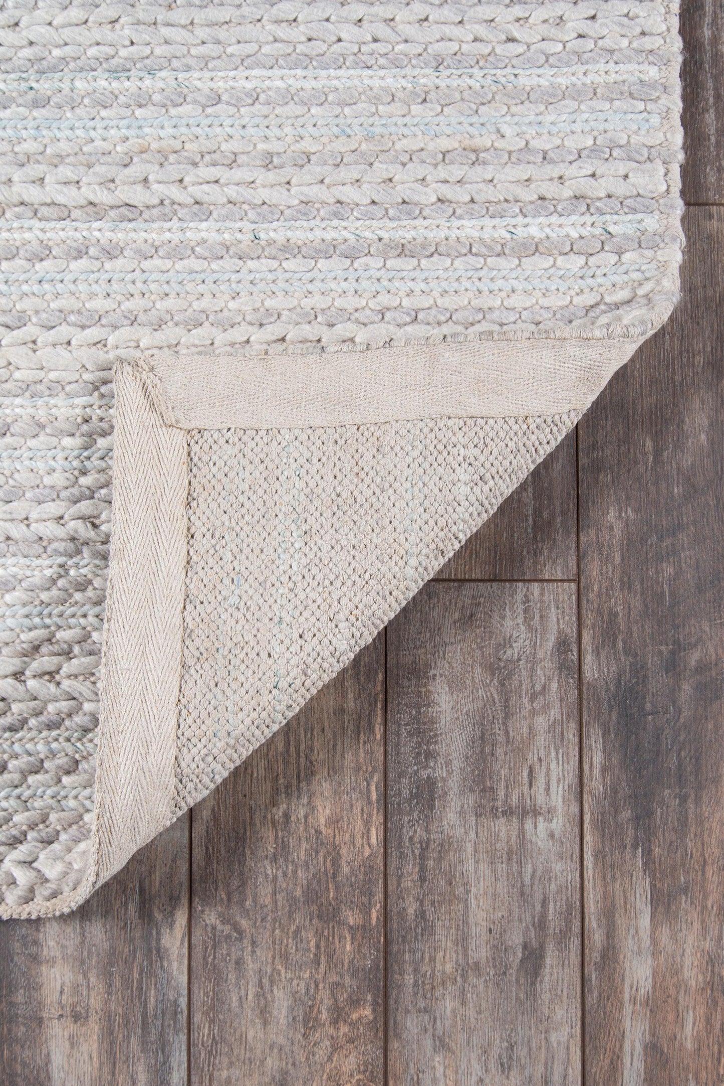 Beige and gray textured woven rug with chunky striped pattern, folded corner exposing underside on dark wood flooring.