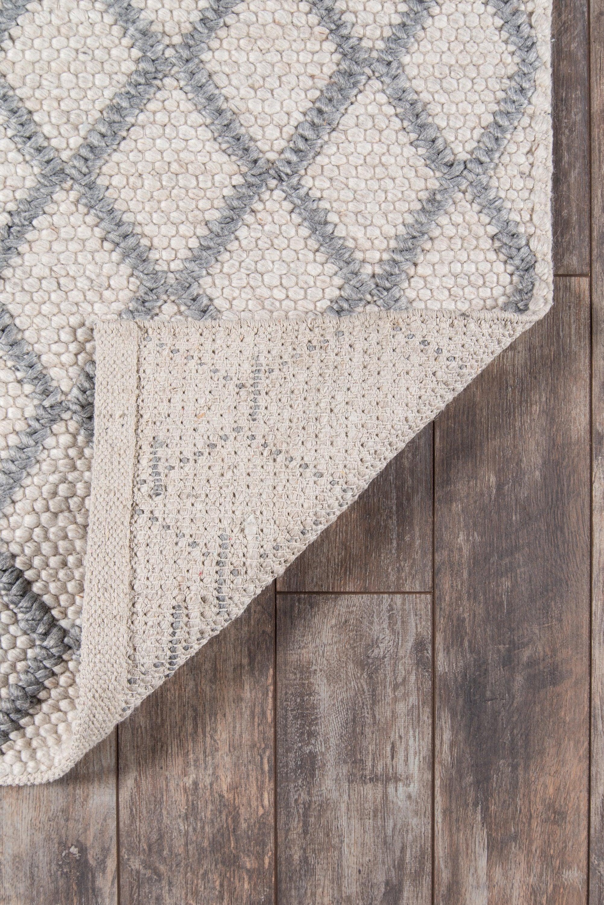 Natural beige rug with grey diamond lattice, loop pile, folded corner showing underside on dark wood floor.