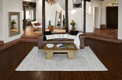 Brown leather sofa with white and black cushions, beige throw, rustic wood coffee table on a light gray rug.