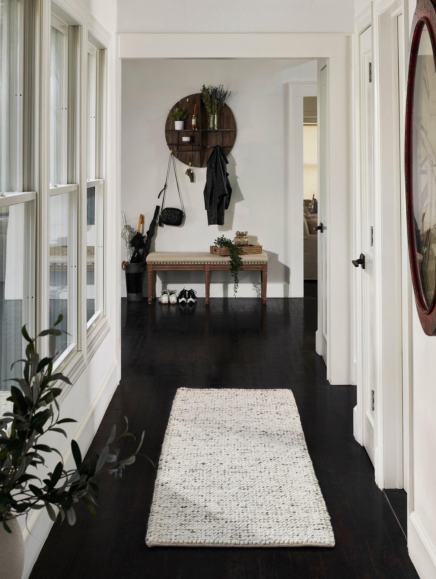 Cream woven hallway runner on dark wood floor with white walls, entry bench, circular wall decor, coat hooks, plants.