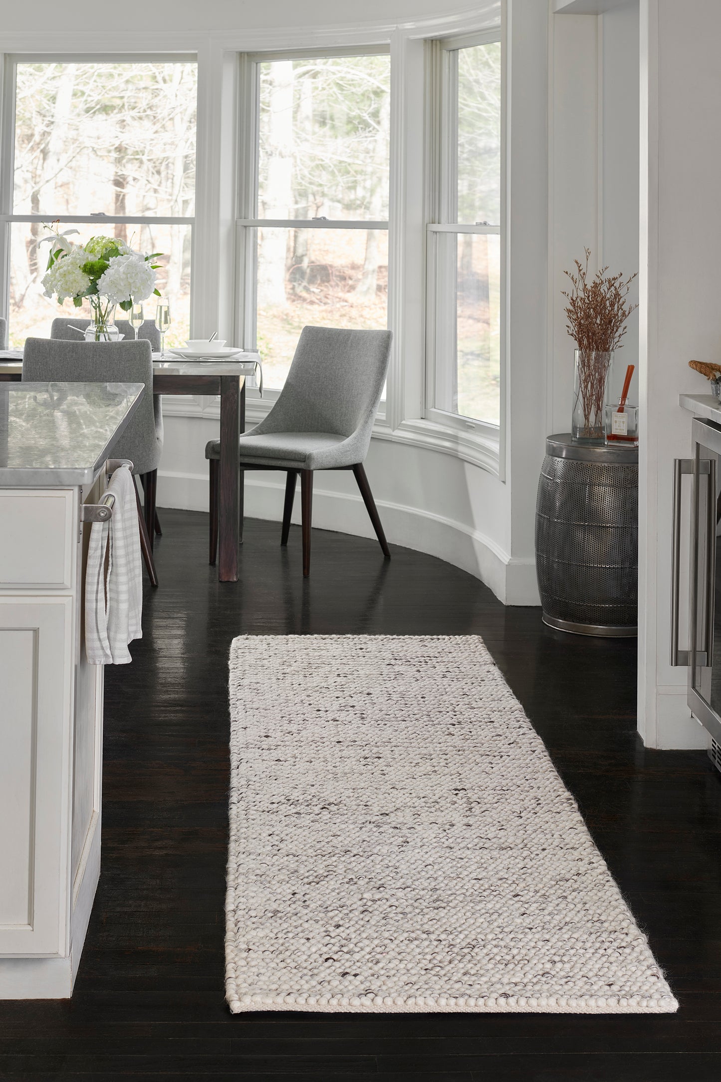 Beige woven runner rug in a modern white kitchen with dark wood floors, bay windows, gray dining chairs.