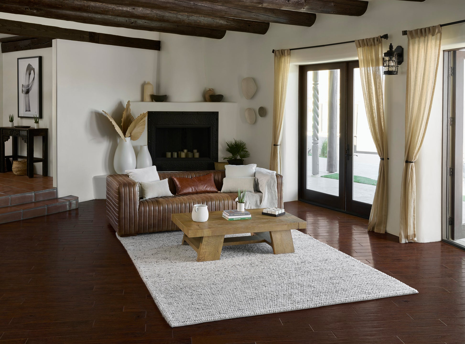 Cozy living room with brown leather sofa, white cushions, wooden coffee table, light rug, exposed beams, beige curtains.