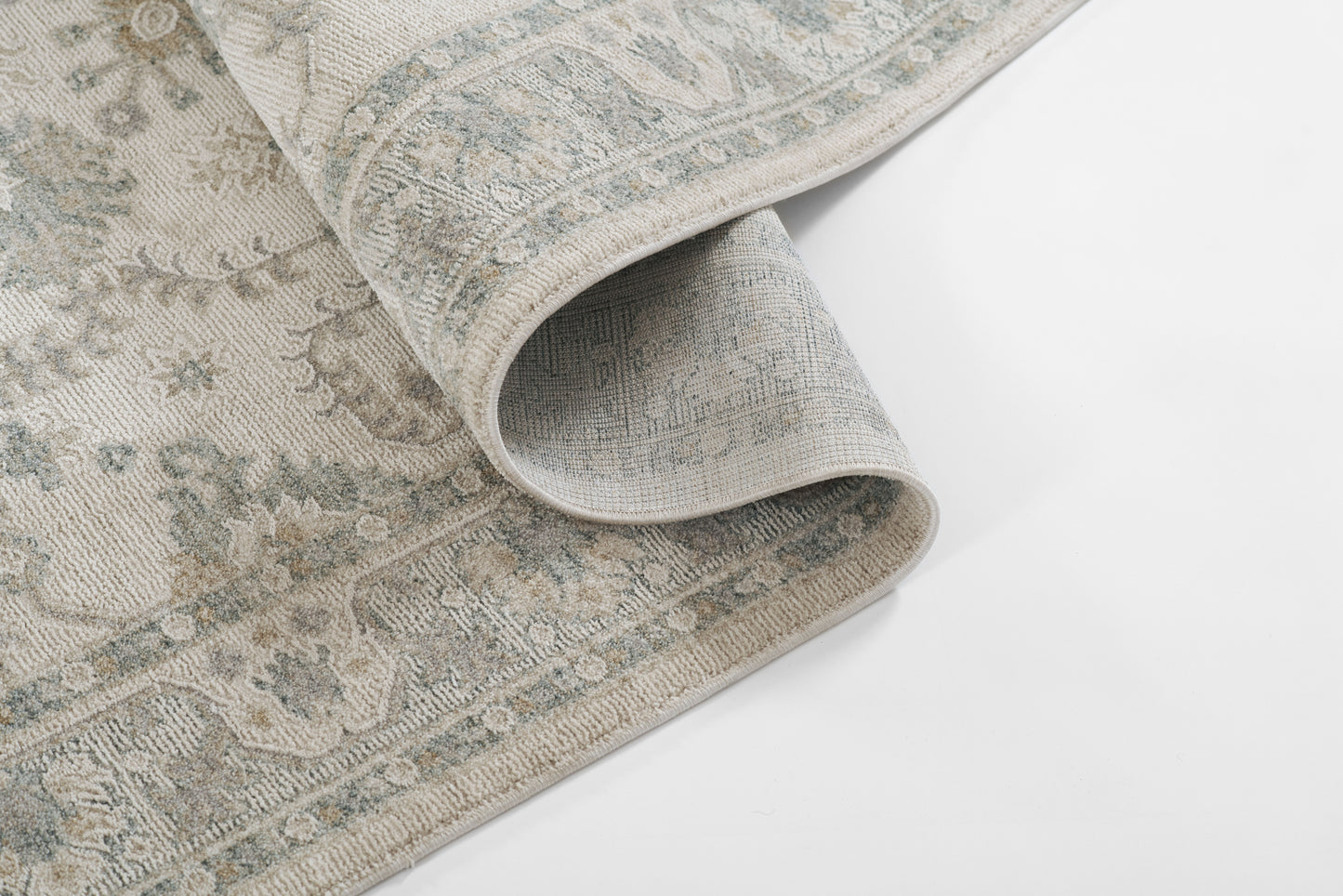 Soft blue-gray rug with antique beige border pattern, woven texture, folded corner showing intricate Persian-inspired design.