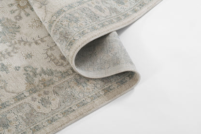 Soft blue-gray rug with antique beige border pattern, woven texture, folded corner showing intricate Persian-inspired design.