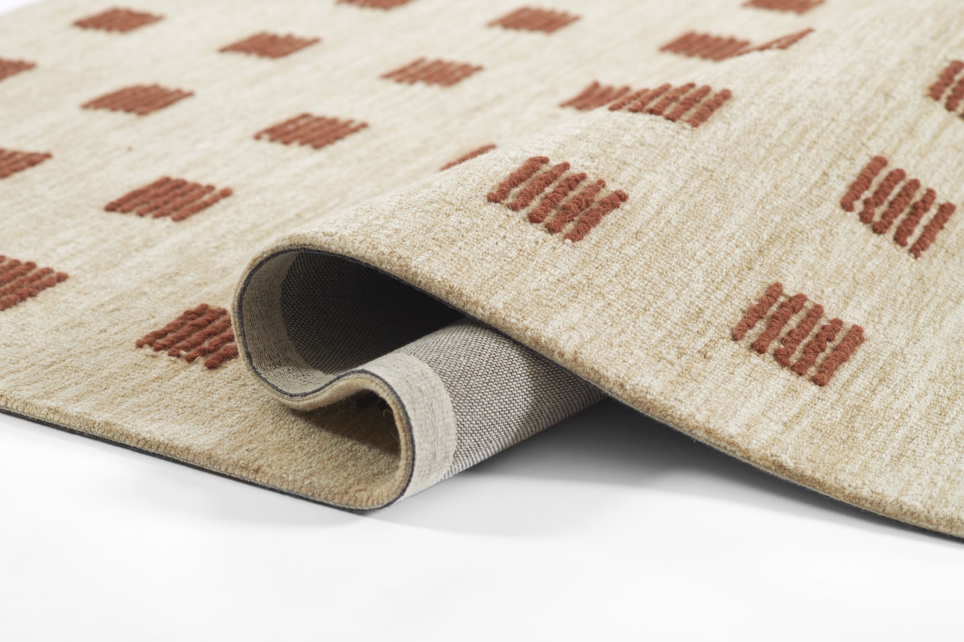 Beige plush area rug with rust-brown diamond motifs; folded corner reveals gray underside.